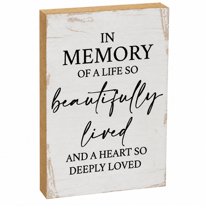 A rectangular wooden memorial sign with a distressed white painted face and natural wood edges. The sign features black text that reads, 'IN MEMORY OF A LIFE SO beautifully lived AND A HEART SO DEEPLY LOVED'.