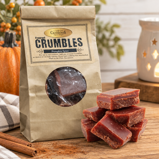 A bag of Crumblelands Spice Company Fragrant Crumbles in Pumpkin Spice flavor stands on a wooden surface next to several pieces of the reddish-brown spiced candy. The scene includes a candle, cinnamon sticks, and a checkered cloth in a warm, styled setting.