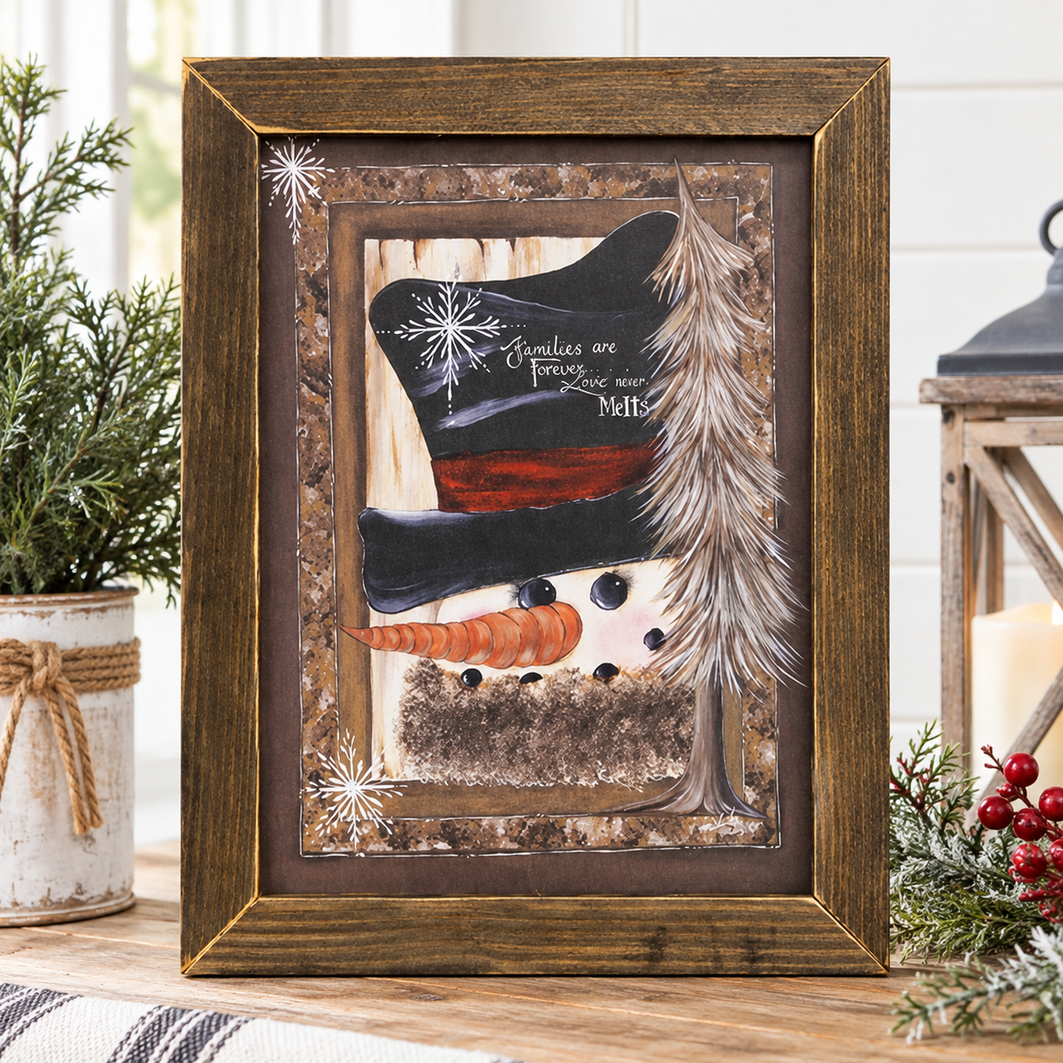 A framed winter-themed art print featuring a snowman face with a carrot nose, a black top hat with snowflakes and the words 'Families are forever. Love our MHS', a feather, and a tree on a rustic wood background, displayed in a wooden frame on a tabletop with holiday greenery and a lantern nearby.