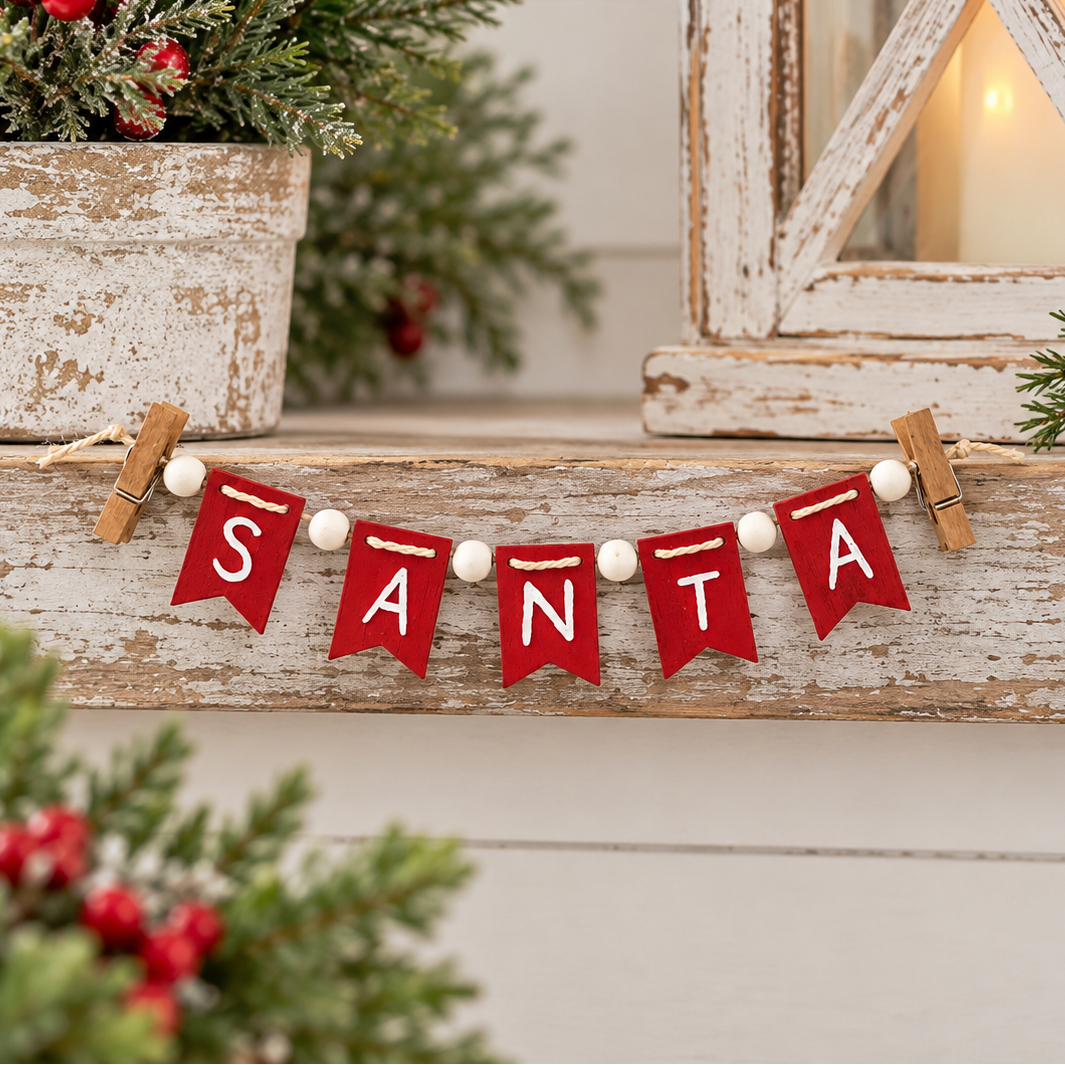 A red felt Christmas garland spelling 'SANTA' with white lettering and small white beads, hung on a rustic wooden shelf with holiday greenery and a lantern in the background.