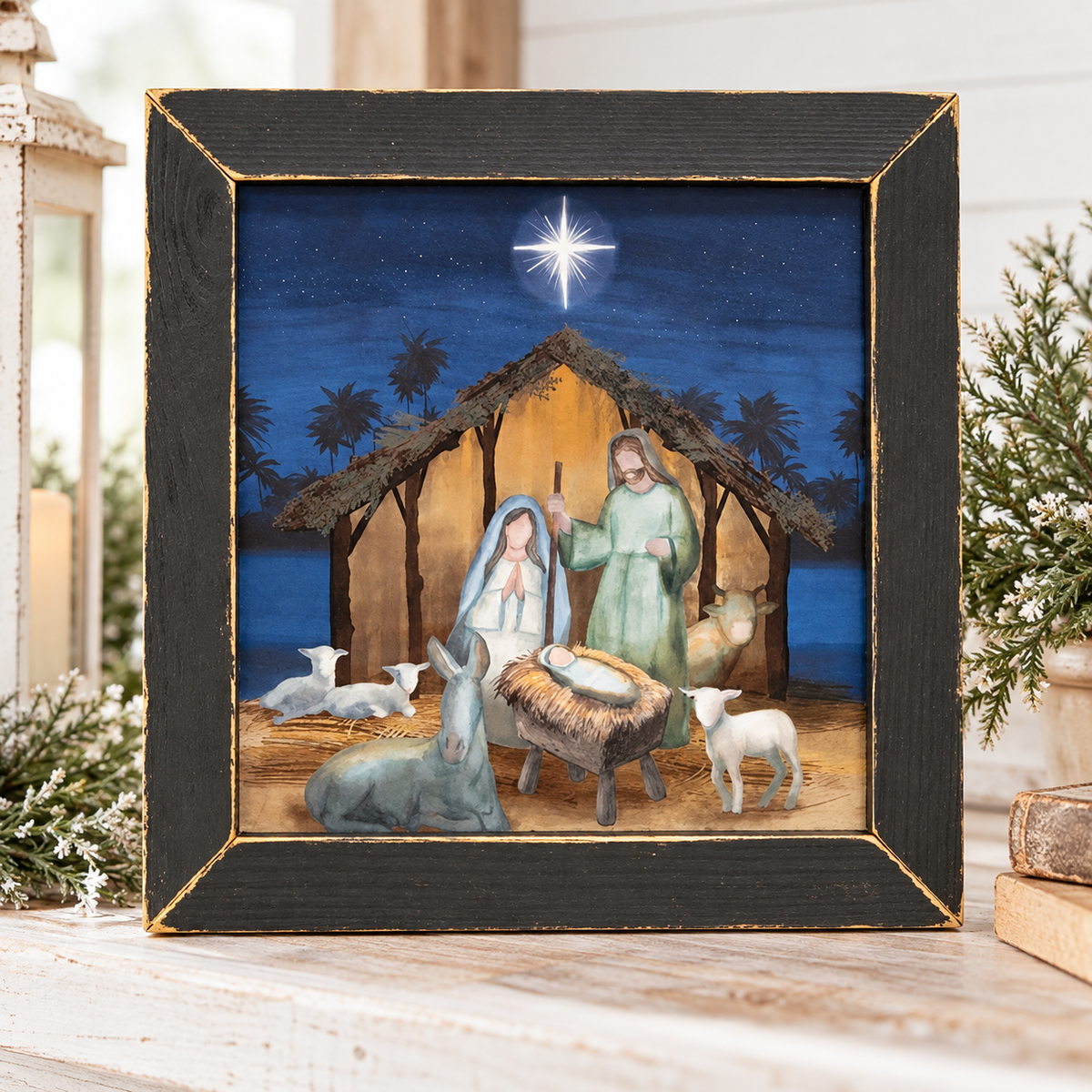A framed Christmas nativity scene print with a black frame and gold trim, displayed on a rustic wooden surface with holiday greenery and lantern props.