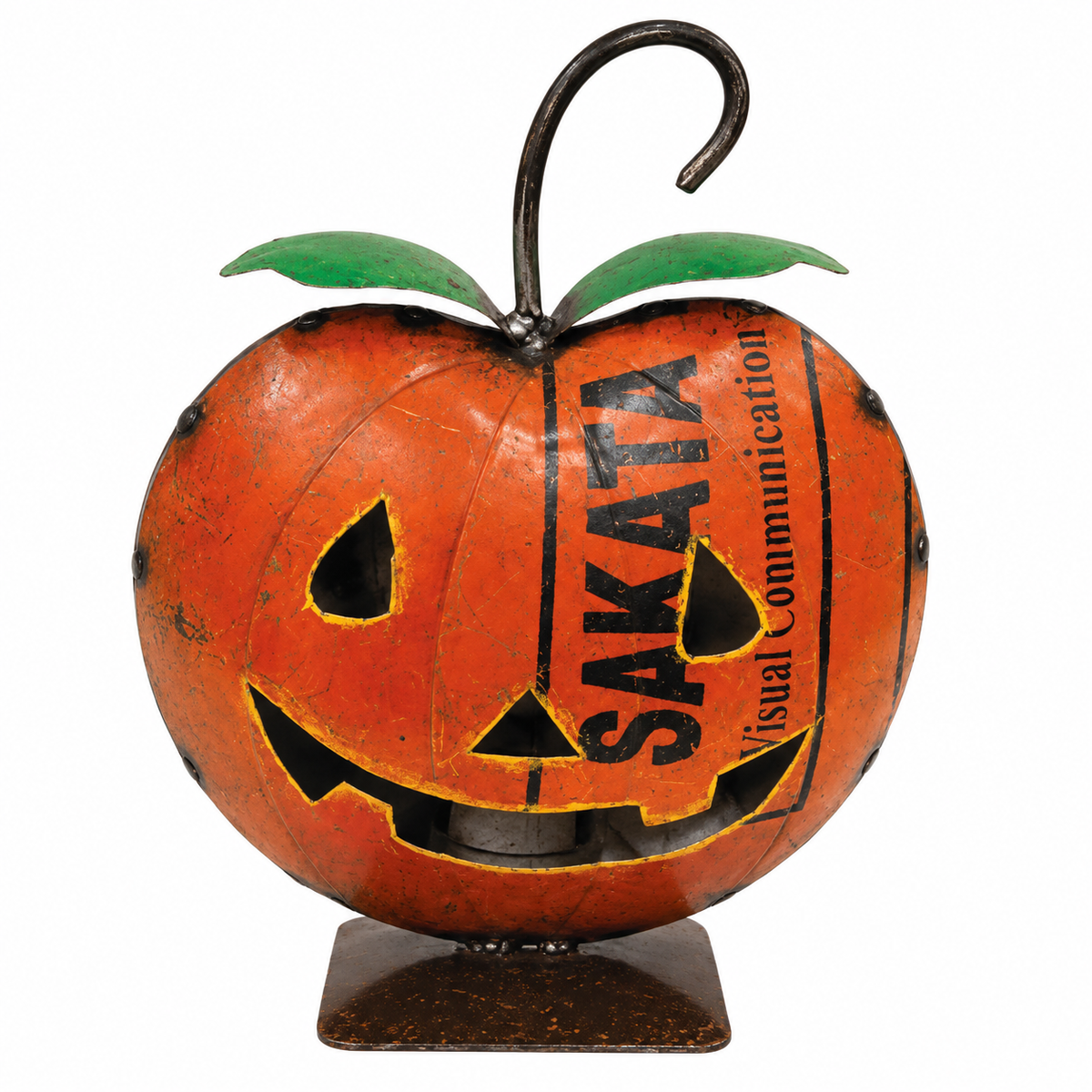 A decorative metal jack-o'-lantern ornament shaped like an apple, with a rusted orange finish, carved facial features, green leaves, a black hanging hook, and the words 'SAKATA Visual Communication' printed vertically on the front, shown on a plain white background.