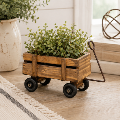A small wooden planter styled like a rustic wagon with black wheels and a metal handle, filled with green leafy plants, placed on a light wood floor in a bright indoor setting.