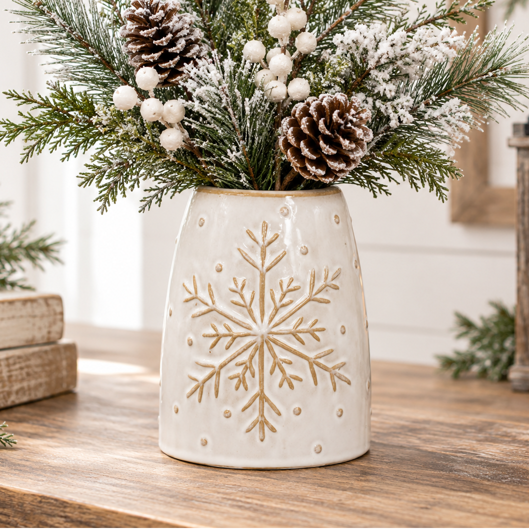 A ceramic holiday vase with a gold snowflake design, filled with faux pine branches, white berries, and pinecones, displayed on a wooden table.