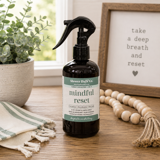 A dark amber spray bottle labeled 'Slower Day Co. mindful reset' sits on a wooden table in a styled home setting with a potted plant, a framed sign, and a tassel bead runner.