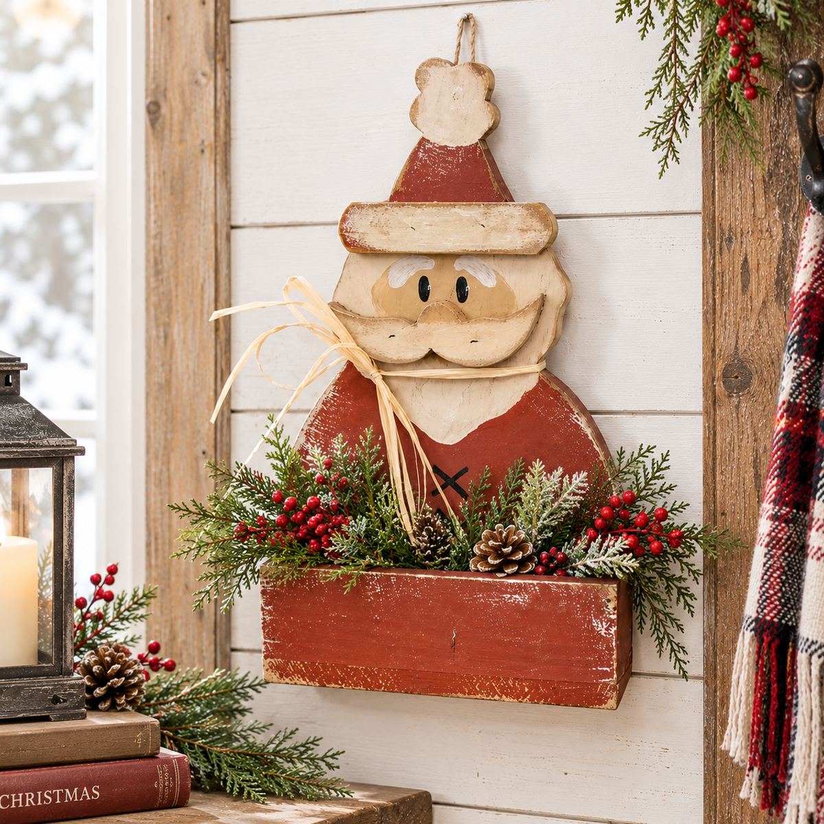 A rustic wooden Santa Claus wall hanging with a red hat and coat, beige beard and face, and a red hanging box filled with green pine branches, red berries, and pinecones, displayed on a white shiplap wall.