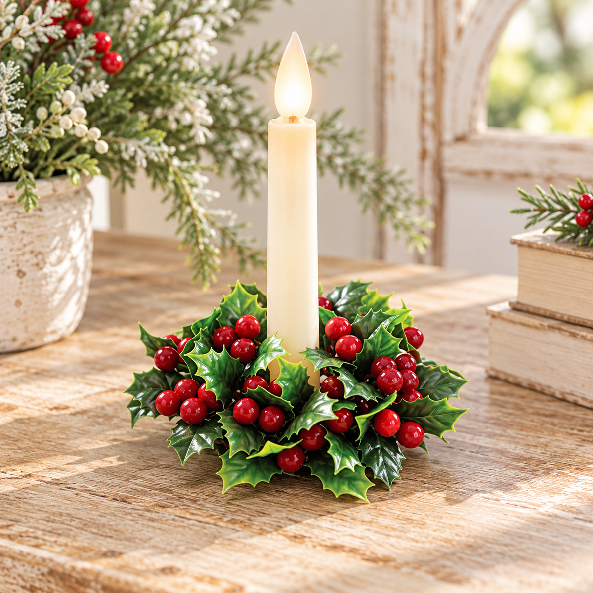 A holiday candle centerpiece with a single cream-colored candle in a circular arrangement of green holly leaves and red berries, set on a rustic wooden table with seasonal decor in the background.