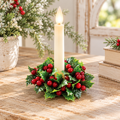 A holiday candle centerpiece with a single cream-colored candle in a circular arrangement of green holly leaves and red berries, set on a rustic wooden table with seasonal decor in the background.