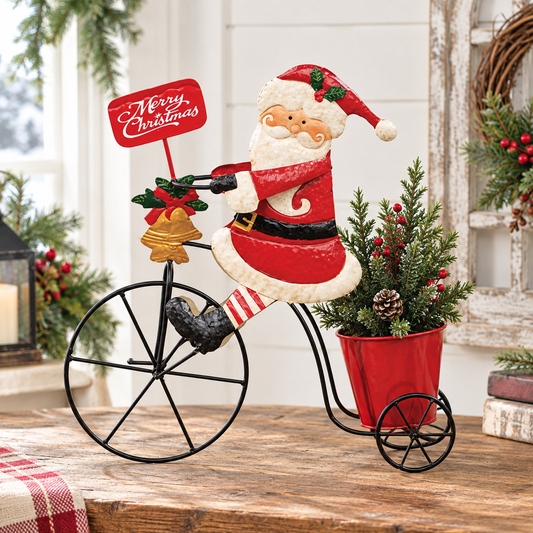 A festive metal decorative figure of Santa Claus riding a black bicycle, with a red sign reading 'Merry Christmas,' greenery, pinecones, and a red planter on a rustic wooden table in a holiday-themed interior setting.
