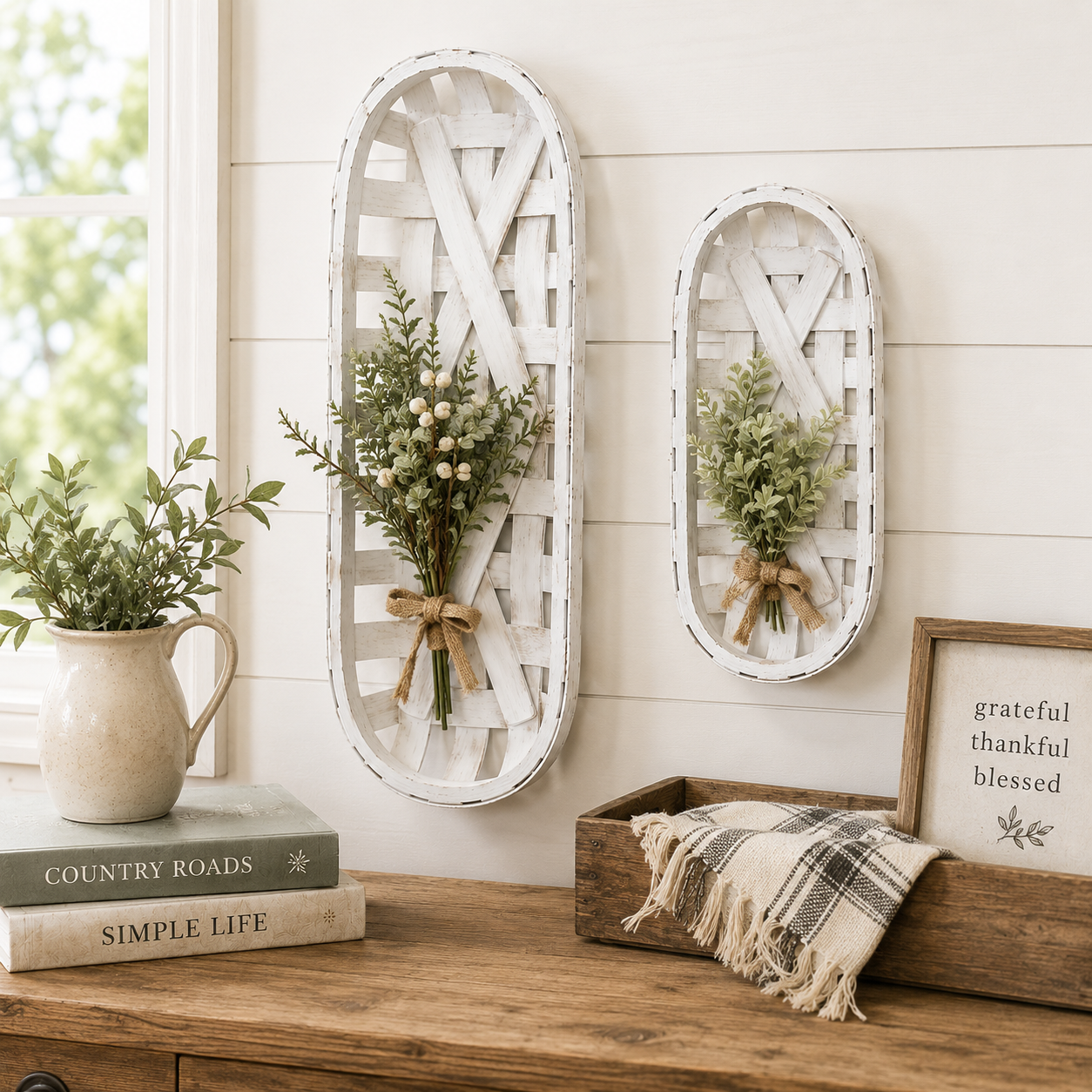 Two white distressed wooden hanging baskets with small green floral arrangements, displayed on a cream shiplap wall above a wooden console table with books, a pitcher, and a framed sign.