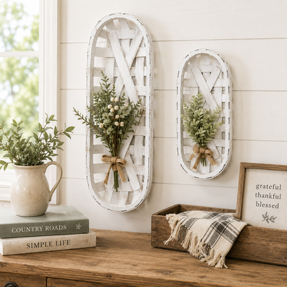 Two white distressed wooden hanging baskets with small green floral arrangements, displayed on a cream shiplap wall above a wooden console table with books, a pitcher, and a framed sign.
