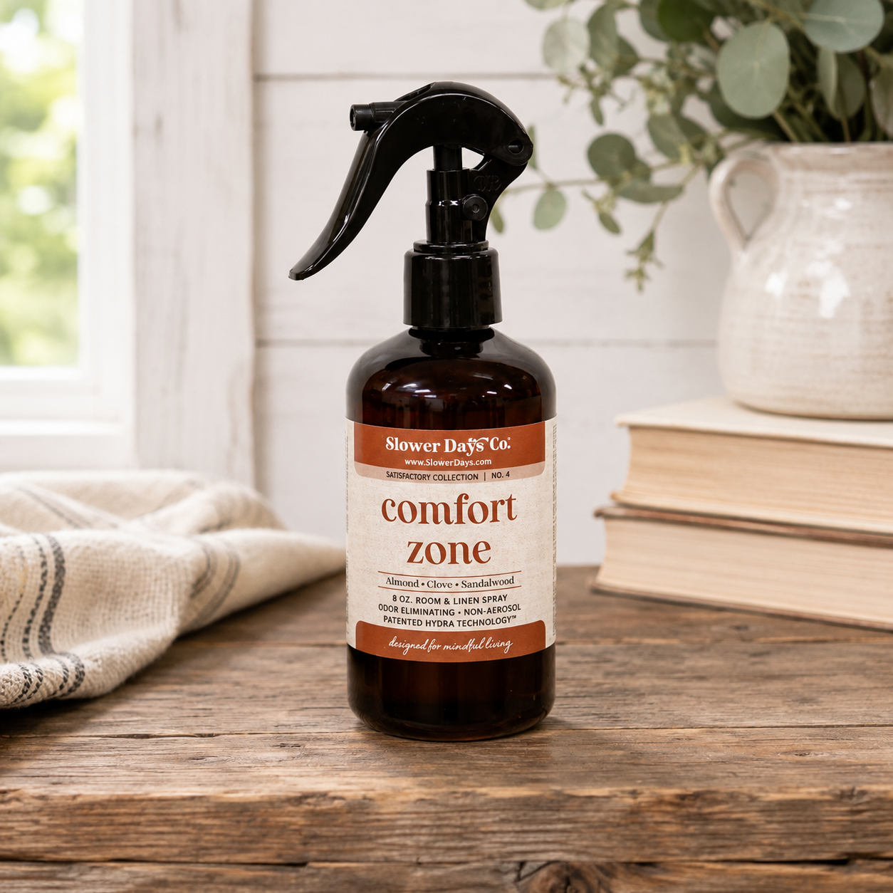 Comfort Zone Room & Linen Spray in an amber glass bottle with black trigger sprayer displayed on a rustic wood table with farmhouse-style décor and soft greenery.