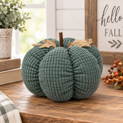 A textured fabric decorative pumpkin in a muted teal green color sits on a wooden table. It features a brown stem and two small gold leaf accents on top, styled in a cozy home setting with a potted plant and a fall-themed sign in the background.