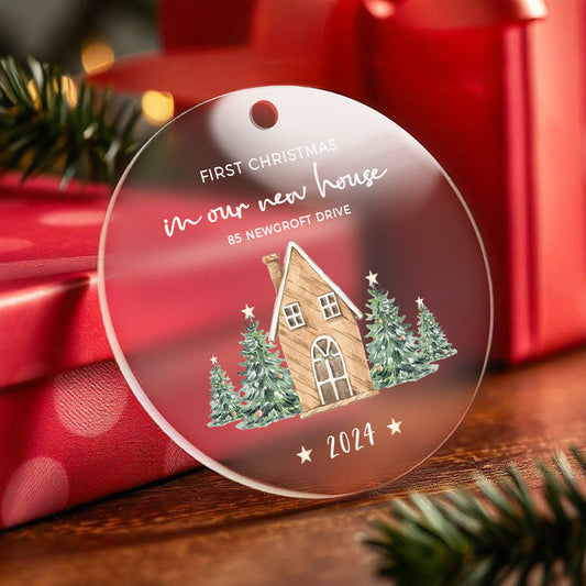 A personalized Christmas ornament featuring a house surrounded by Christmas trees with the text "First Christmas in our new home." The ornament can be customized with the customer’s address and year for a special, personalized touch.