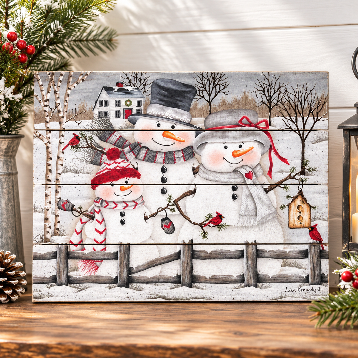 A rectangular winter-themed decorative sign or canvas showing two snowmen, a small snowman, bare trees, a house, fence, and red birds, displayed on a wooden surface with holiday greenery and a lantern around it.