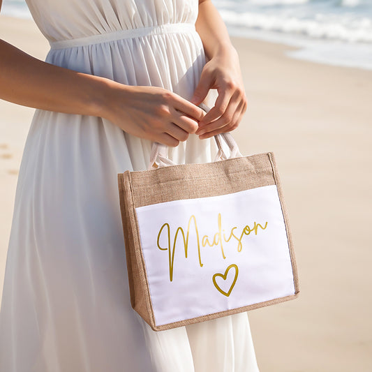 A woman standing on the beach holding a personalized jute tote bag, showcasing its spacious design, durable linen fabric, and convenient front pocket for easy access to essentials.