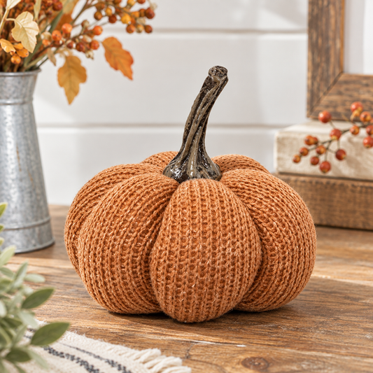 A decorative knitted pumpkin in a warm rust-orange color with a dark brown faux-stem, displayed on a wooden tabletop with fall-themed rustic decor in the background.