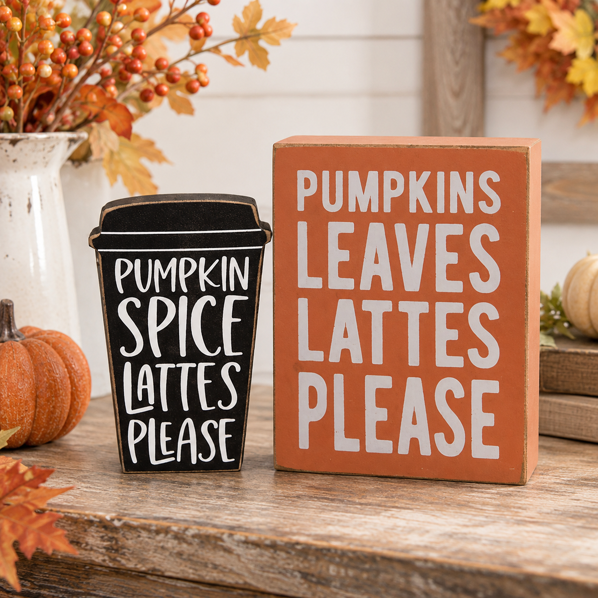 Two fall-themed decorative signs on a rustic wooden table: a black coffee cup sign reading 'PUMPKIN SPICE LATTES PLEASE' and an orange block sign reading 'PUMPKINS LEAVES LATTES PLEASE', styled with autumn leaves, berries, and small pumpkins.