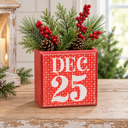A square red decorative planter with white polka dots and the words 'DEC 25' in large white letters, filled with pine branches, pinecones, and red berries, set on a rustic wooden surface.