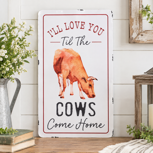 A rectangular white decorative sign with a red border and a watercolor-style brown cow in the center, surrounded by home decor props including flowers, books, and a wooden table against a white shiplap wall.