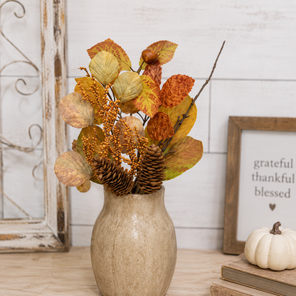 Autumn artificial floral arrangement in a beige ceramic vase, featuring orange and yellow leaves, pinecones, and small orange berries, styled on a tabletop with a framed sign and a small pumpkin in the background.