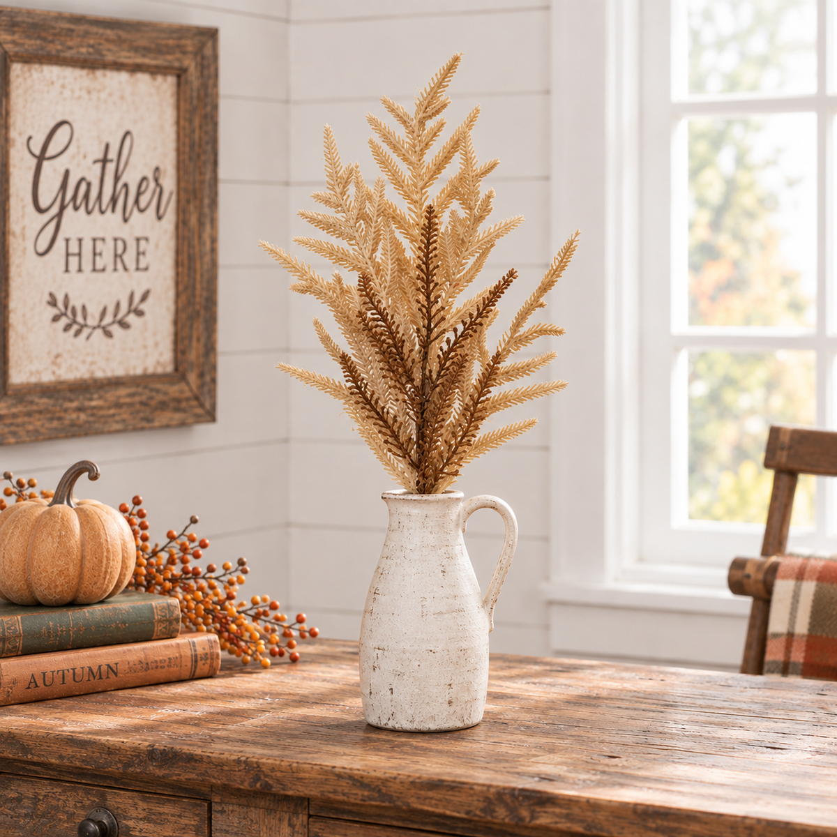 A decorative artificial fern arrangement in a white ceramic pitcher vase, styled on a rustic wooden table with autumn books, a small pumpkin, and berries in a bright room with a window and framed sign.