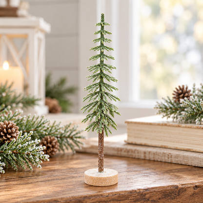 A small decorative evergreen tree ornament with a glittery green textured body and a brown trunk on a round cork base, displayed on a wooden table with holiday decor in a softly lit indoor setting.