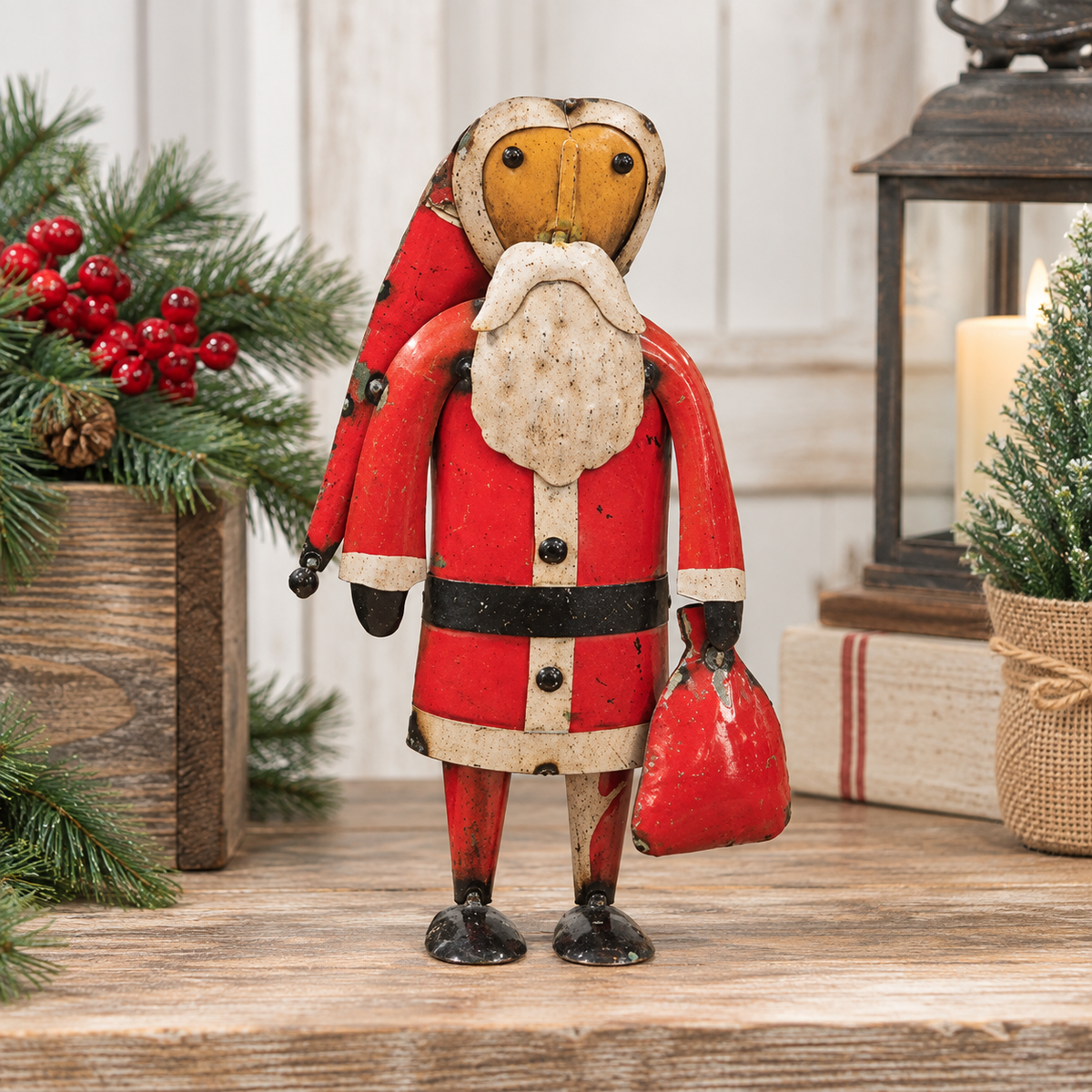 A rustic hand-painted wooden Santa Claus figurine standing on a wooden surface, surrounded by holiday decorations including pine branches with red berries, wrapped gifts, and a lantern.