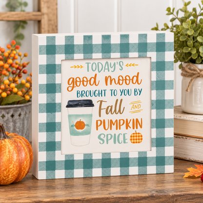 A square decorative sign with a white and teal gingham border and autumn-themed text reading 'TODAY'S good mood BROUGHT TO YOU BY Fall AND PUMPKIN SPICE,' shown on a wooden surface with pumpkins, fall leaves, and plants around it.