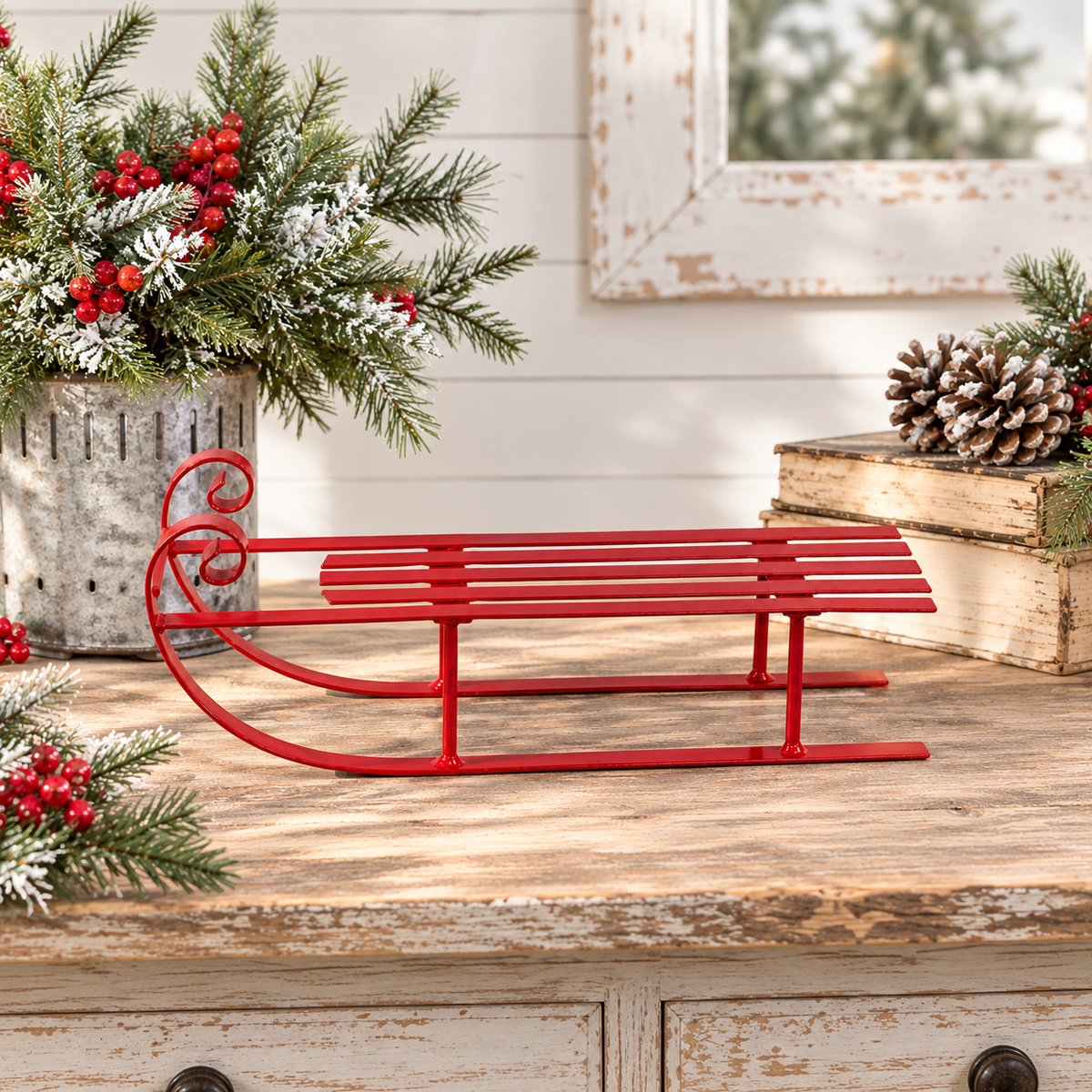 A red metal sleigh-shaped decorative object sits on a rustic wooden surface, styled with holiday greenery, red berries, books, and a distressed white wall in the background.