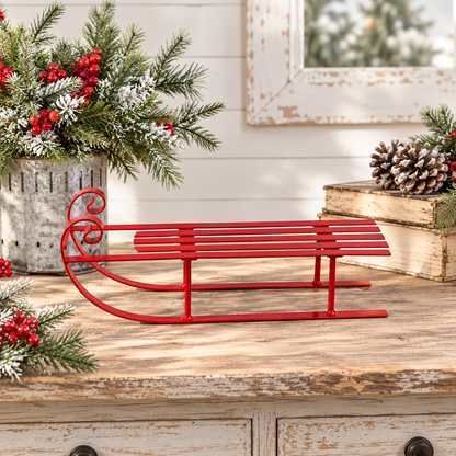 A red metal sleigh-shaped decorative object sits on a rustic wooden surface, styled with holiday greenery, red berries, books, and a distressed white wall in the background.
