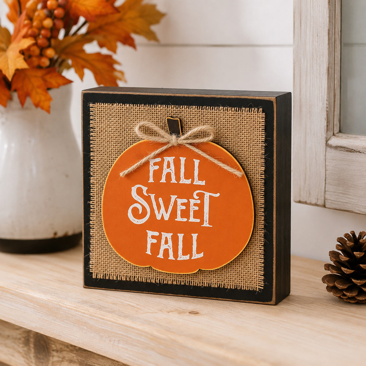 A square decorative sign with a black frame and burlap-style front, featuring an orange pumpkin shape with the words “FALL SWEET FALL” in white lettering and a small twine bow. The sign sits on a wooden surface with fall decor in the background.