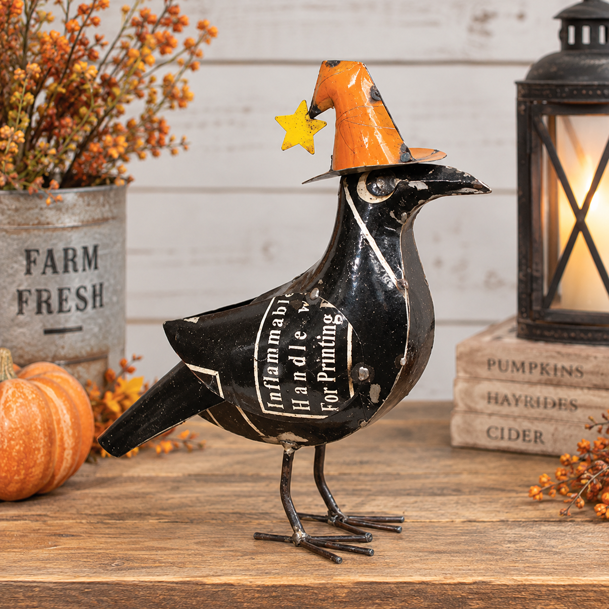 A decorative black metal crow figurine wearing an orange witch hat with a yellow star, displayed on a wooden surface with fall-themed props including a small pumpkin, books, and a lantern.