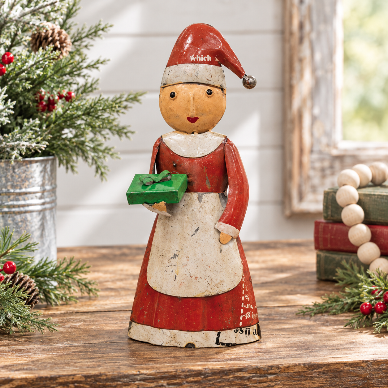 A rustic wooden Santa figurine with a distressed red and cream painted finish, holding a small green gift box, displayed on a wooden table with holiday decorations.