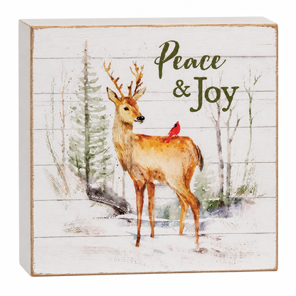 Square decorative wall sign with a watercolor-style deer standing in a snowy winter scene, a small red bird on the deer's back, and the words 'Peace & Joy' on a distressed white wood plank background.