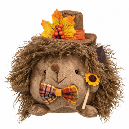 A plush hedgehog decoration with shaggy brown faux fur, wearing a brown hat with an orange and yellow autumn leaf, a plaid bow tie, and holding a small sunflower on a stick, shown on a plain white background.
