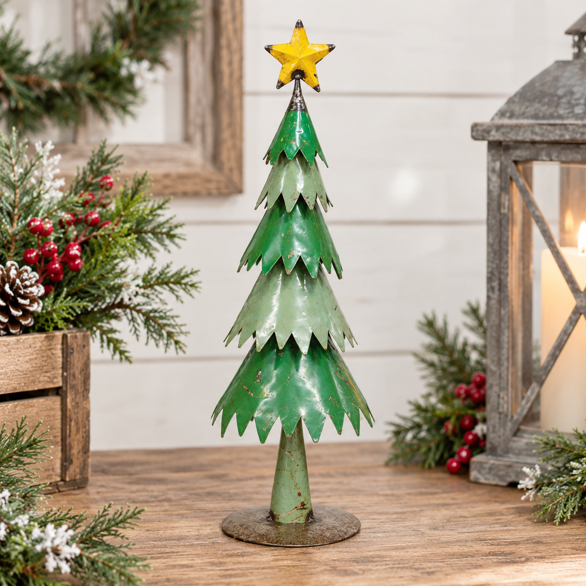 A small decorative metal Christmas tree in distressed green with a yellow star topper, displayed on a wooden surface with holiday greenery and a lantern in the background.