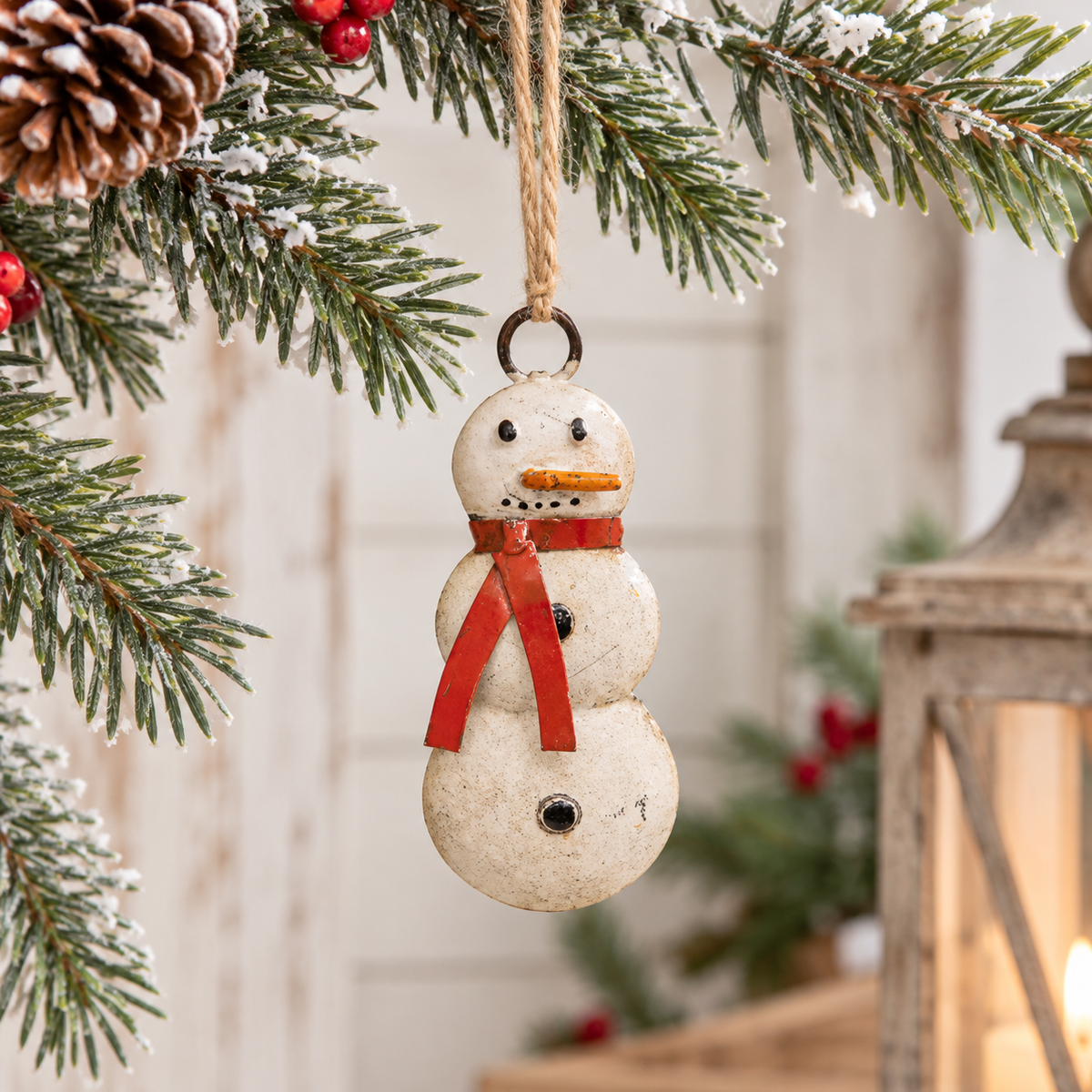 A small snowman Christmas ornament hanging from a pine tree branch. The ornament has a distressed off-white finish with a red scarf, black eyes and buttons, and a small orange carrot nose.