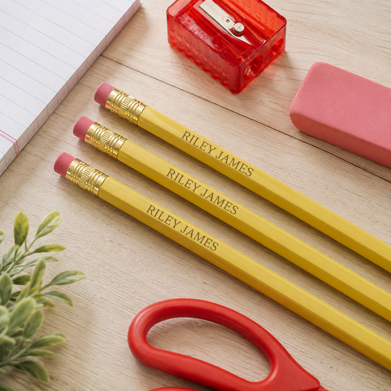Three personalized yellow pencils with pink erasers and gold ferrules are arranged on a light wood desk alongside a red pencil sharpener, pink eraser, scissors, lined paper, and a small plant.