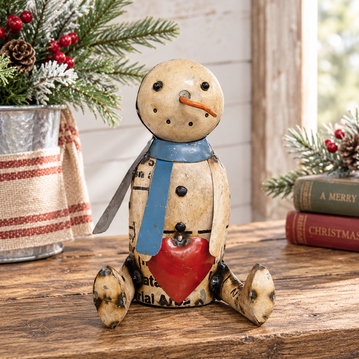 A rustic ceramic snowman figurine sitting on a wooden table, decorated with a blue scarf, red heart, and black printed text details, surrounded by holiday decorations.