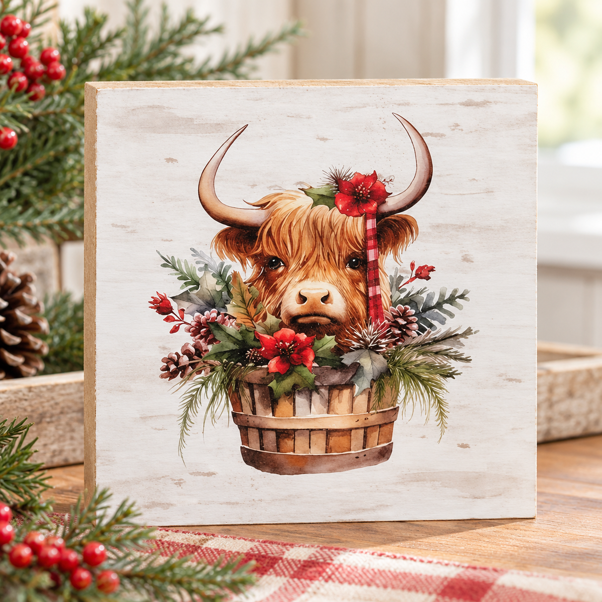 A square decorative wooden sign featuring a watercolor illustration of a Highland cow in a basket with red berries and evergreen branches, styled on a wooden surface with holiday greenery and red berries in the foreground.