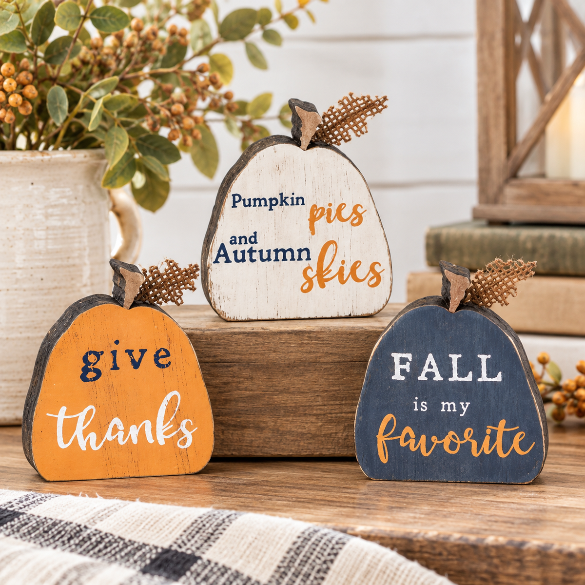 Three decorative autumn wooden sign pumpkins displayed on a rustic wooden surface. One orange pumpkin reads 'give thanks' in navy and white lettering.