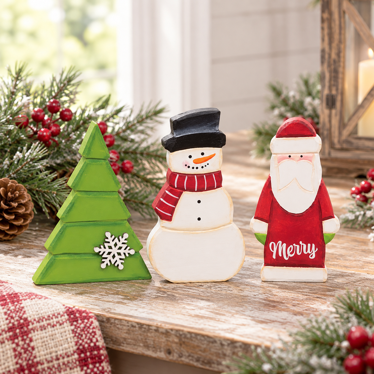Three hand-painted wooden Christmas decorations on a rustic wooden table: a green Christmas tree with a white snowflake, a snowman in a black top hat and red striped scarf, and a Santa Claus figure with 'Merry' written on his chest. The scene is decorated with pine branches, red berries, and a pinecone.