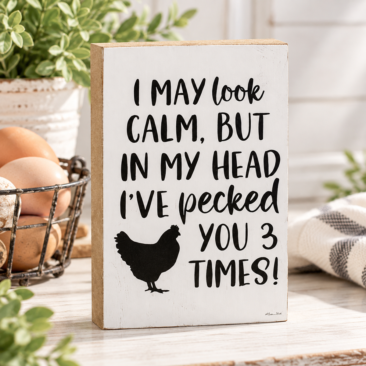 A rectangular white wooden sign with a natural wood edge, featuring black text that reads 'I MAY look CALM, BUT IN MY HEAD I'VE pecked YOU 3 TIMES!' and a black chicken silhouette. The sign is styled on a rustic table with eggs, greenery, and a checkered cloth in the background.