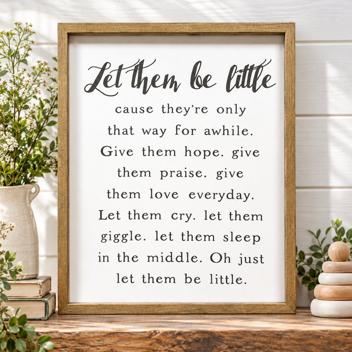 A framed typographic wall sign with the phrase “Let them be little” in black script and serif text on a white background, set in a rustic wood frame. The sign is displayed on a wooden surface with decorative plants, books, and stacked wooden beads around it.