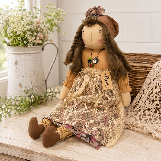 Primitive country rag doll named Rosie with embroidered facial features, braided brown hair, brown fabric hat, floral dress, ivory lace apron, and button necklace for farmhouse and rustic home décor.